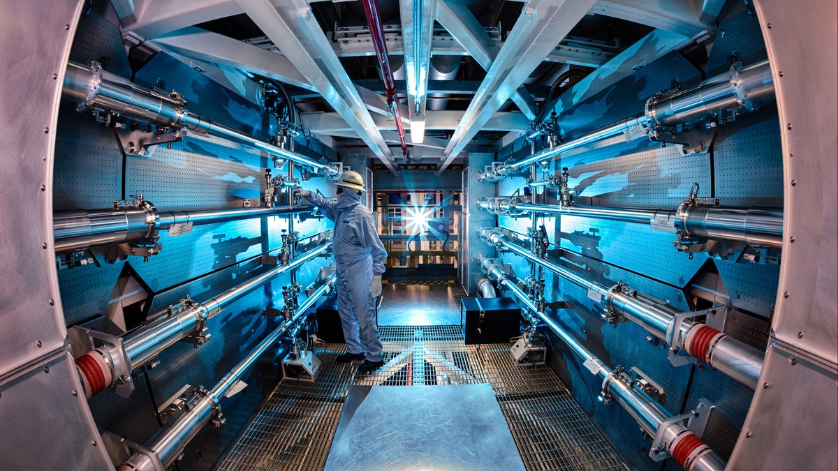 A man in a white jumpsuit adjusts something in a sort of cylindrical room with blueish lights. Metallic tubes adorn both sides of the room.